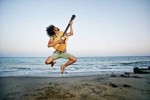 Fotografi Mixed Race man playing guitar and jumping at beach, Peathegee Inc, (40 x 26.7 cm)